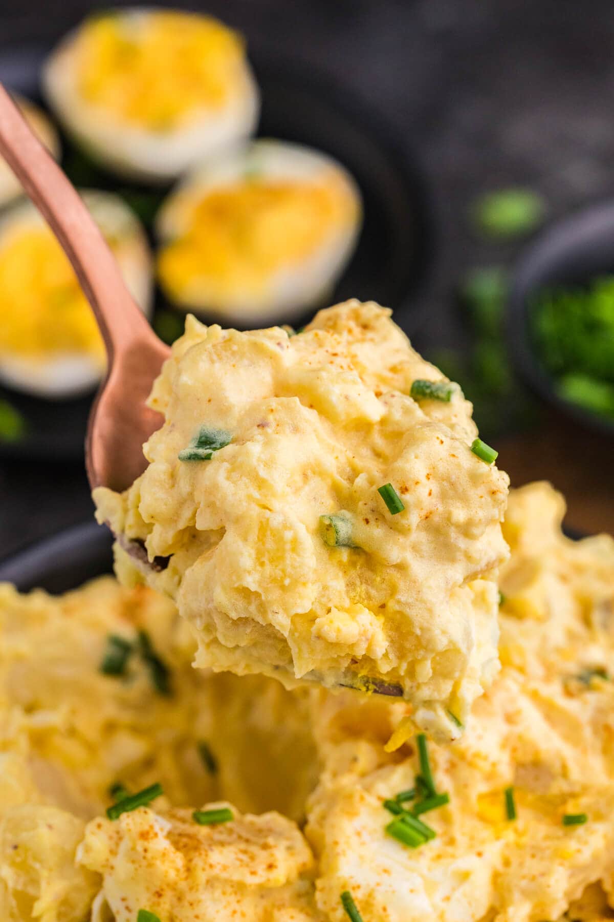 Spoonful of creamy Deviled Egg Potato Salad with chunks of potato and egg, topped with paprika and chopped chives.
