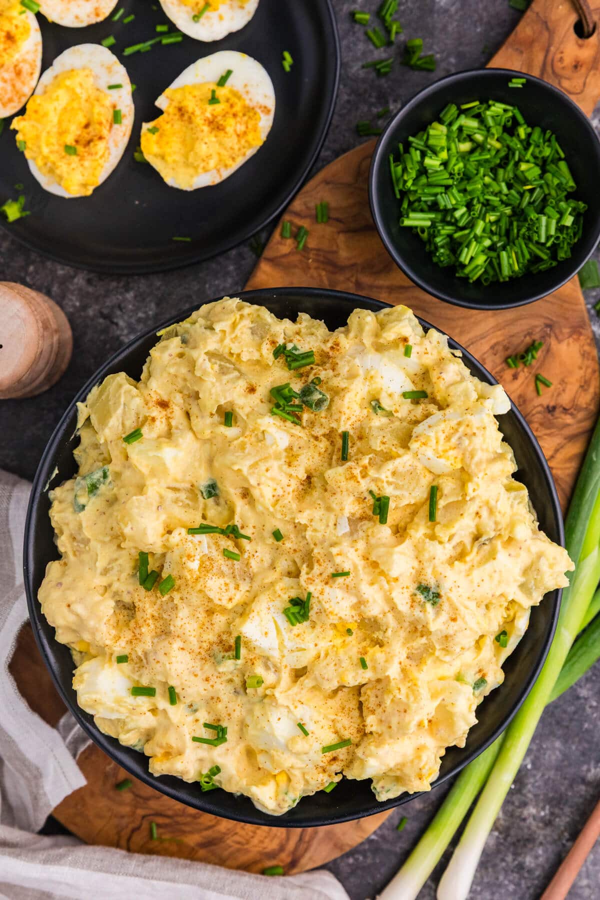 Creamy Deviled Egg Potato Salad with chopped eggs, paprika, and chives in a black bowl, with deviled eggs and green onions nearby.