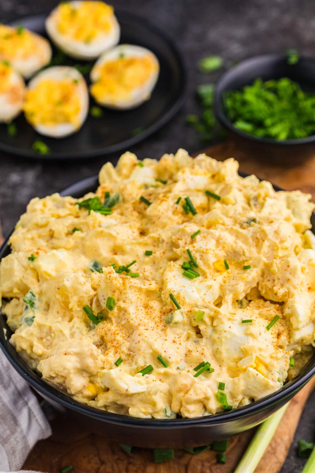 Close-up of creamy Deviled Egg Potato Salad with chopped eggs, paprika, and chives in a black bowl.