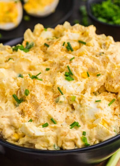 Creamy Deviled Egg Potato Salad topped with paprika and chopped chives, with deviled eggs and green onions in the background.