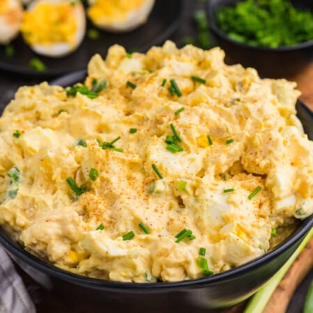 Creamy Deviled Egg Potato Salad topped with paprika and chopped chives, with deviled eggs and green onions in the background.