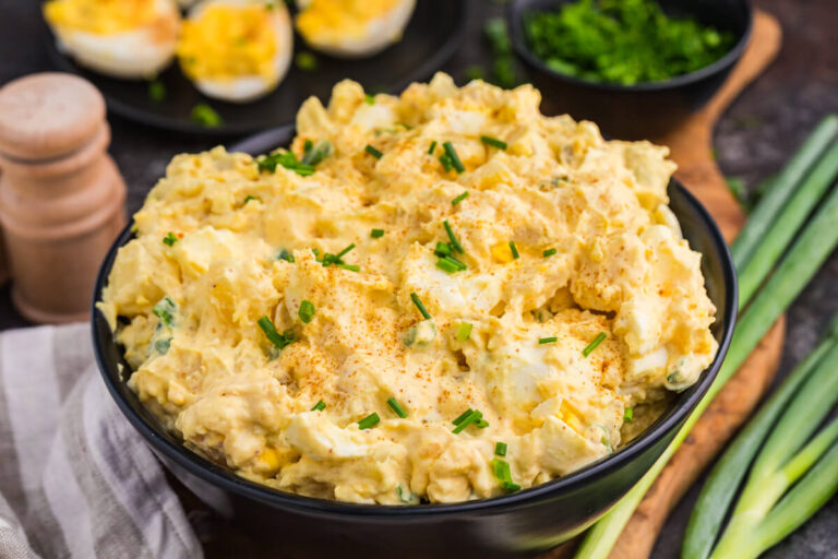 Creamy Deviled Egg Potato Salad topped with paprika and chopped chives, with deviled eggs and green onions in the background.