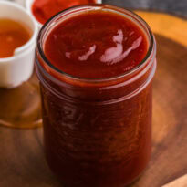 Jar of homemade Honey Sriracha BBQ Sauce with honey and Sriracha in the background.