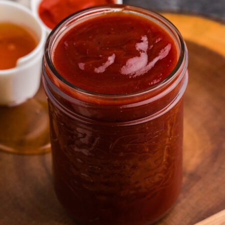 Jar of homemade Honey Sriracha BBQ Sauce with honey and Sriracha in the background.