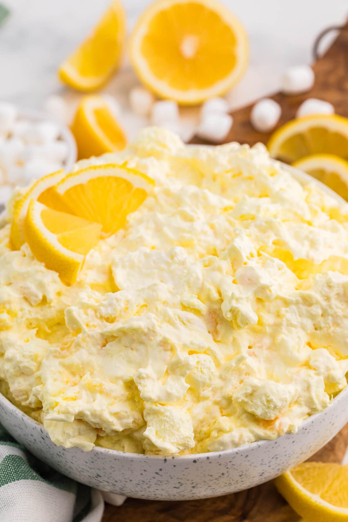 Close-up of creamy Lemon Fluff dessert salad topped with lemon slices, with mini marshmallows and fresh lemons in the background.