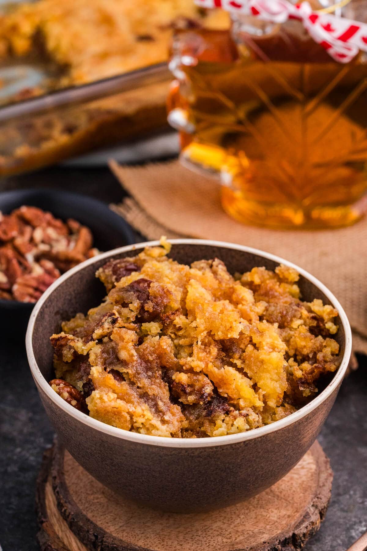 Bowl of Maple Syrup Dump Cake with pecans and gooey maple filling, with a maple syrup bottle in the background.