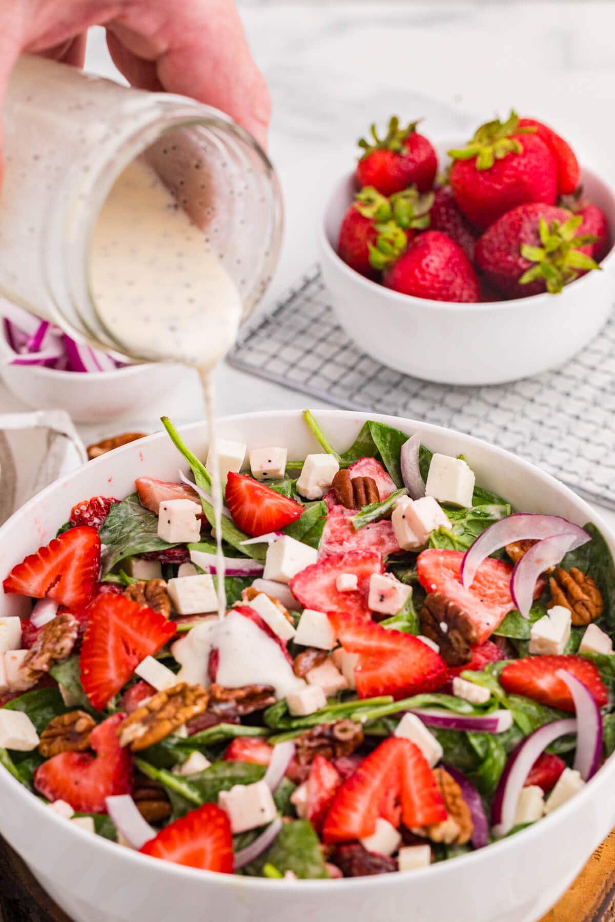 Creamy poppyseed dressing being poured over strawberry spinach salad with feta cheese, toasted pecans, and red onion.