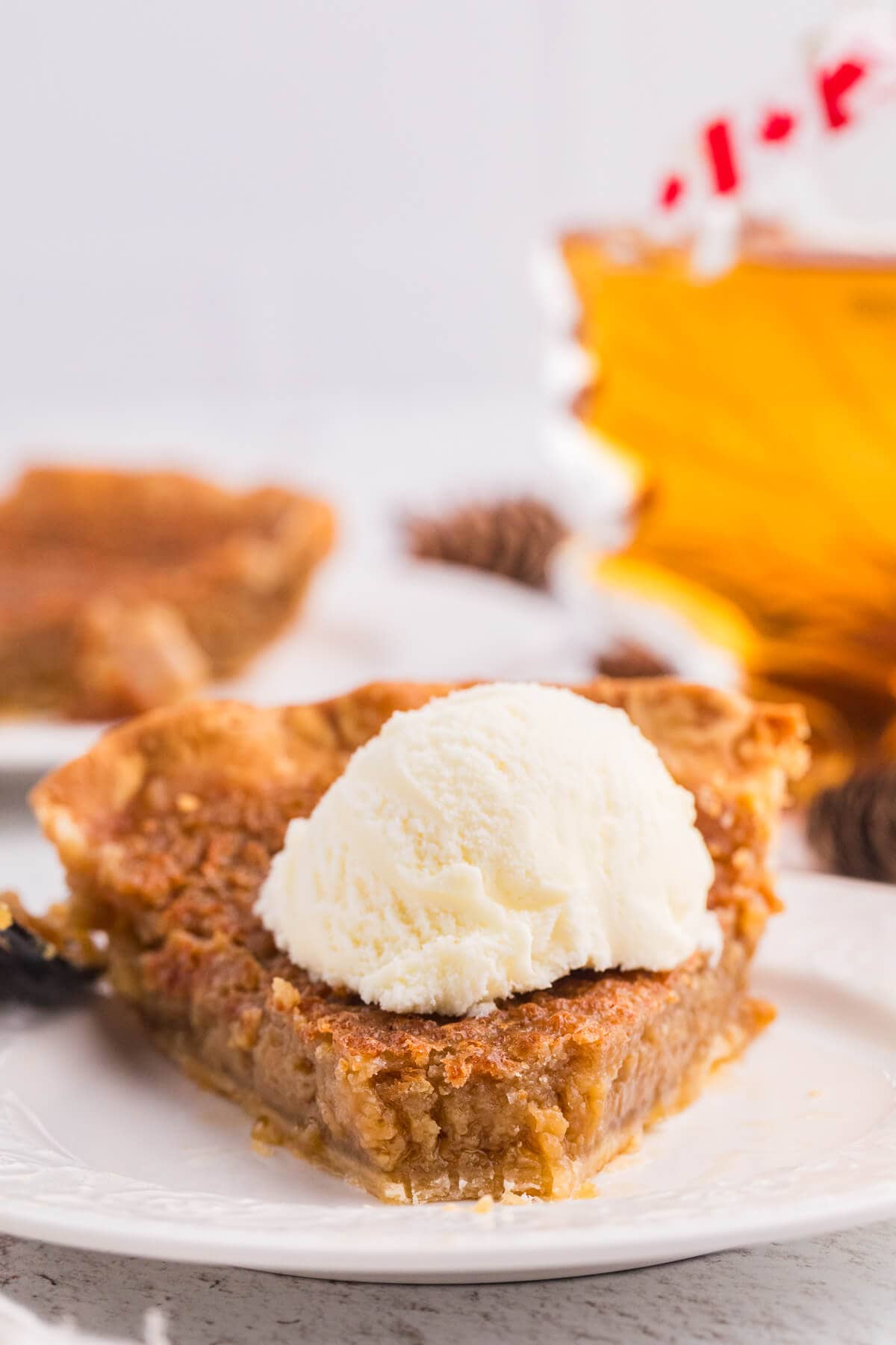 Close-up of Maple Syrup Pie topped with a scoop of vanilla ice cream, with maple syrup and another slice of pie in the background.