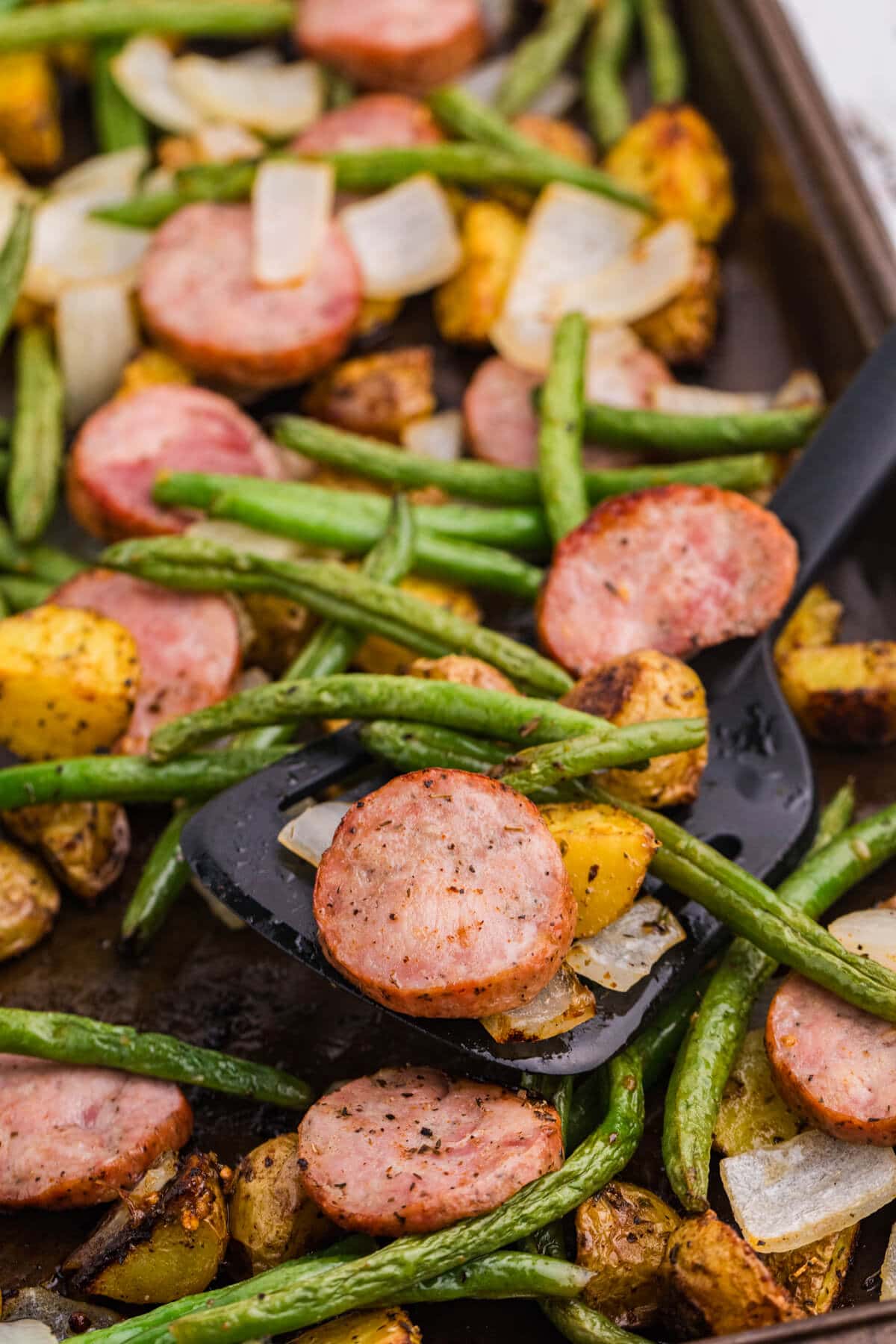 Close-up of sheet pan sausage, potatoes and green beans with roasted kielbasa, baby potatoes, green beans, and onion being served from the pan.