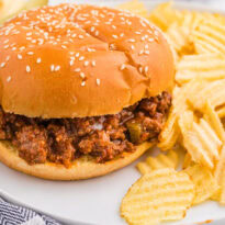 Homemade Sloppy Joe sandwich with saucy ground beef on a sesame seed bun, served with ridged potato chips and pickles.