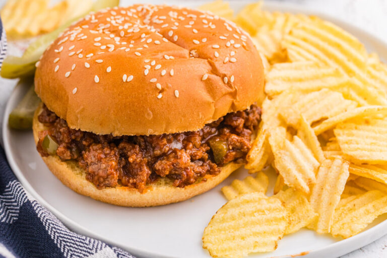 Homemade Sloppy Joe sandwich with saucy ground beef on a sesame seed bun, served with ridged potato chips and pickles.