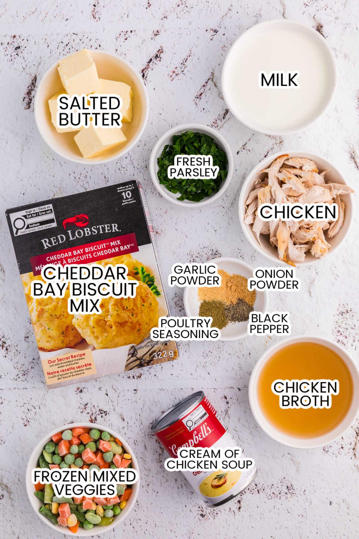 Ingredients for TikTok Chicken Cobbler including shredded chicken, frozen mixed vegetables, Cheddar Bay biscuit mix, cream of chicken soup, chicken broth, milk, butter, seasonings, and parsley.
