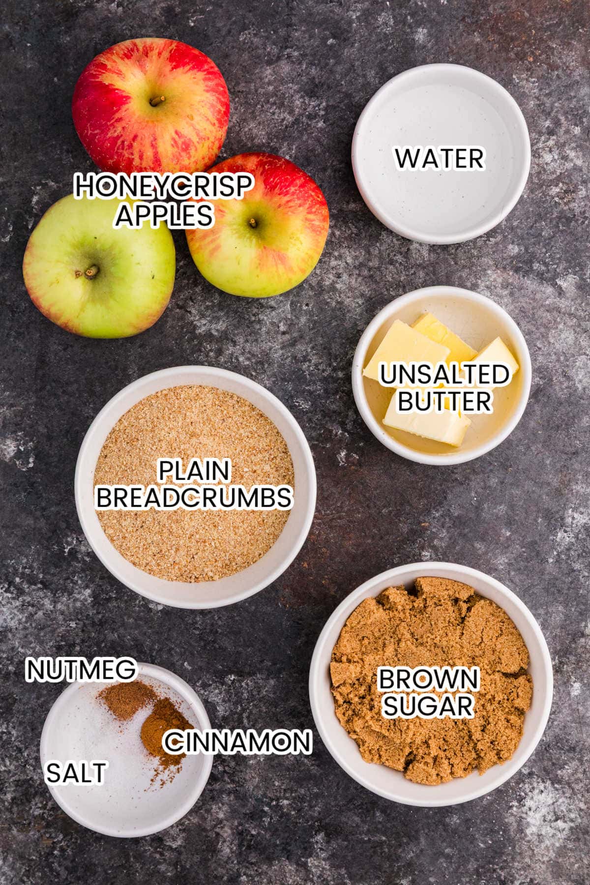 Ingredients for apple brown betty laid out in bowls and labeled: Honeycrisp apples, water, unsalted butter, plain breadcrumbs, brown sugar, nutmeg, cinnamon, and salt.