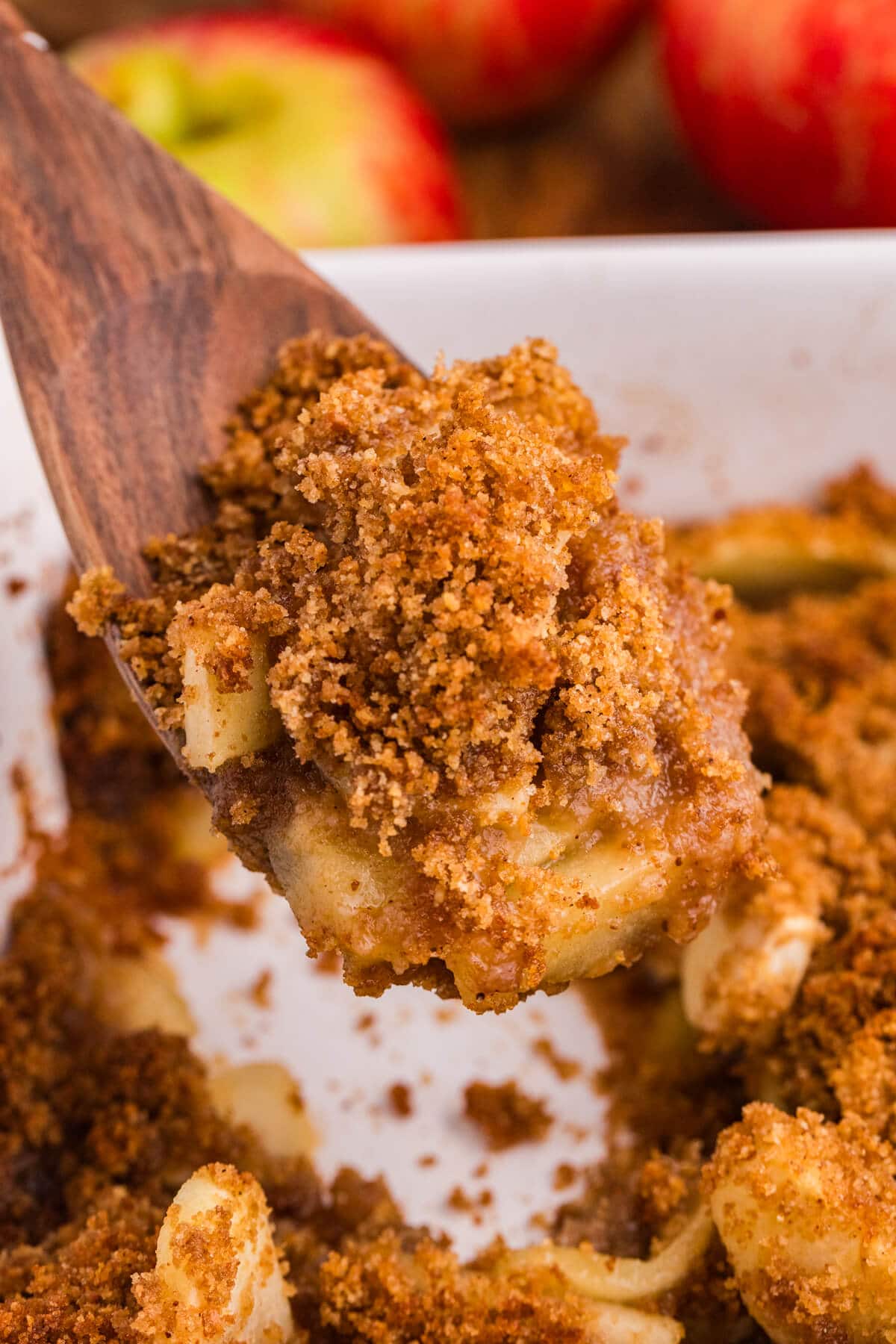 Wooden spatula lifting a scoop of apple brown betty with tender apple slices and a golden brown breadcrumb topping from a white baking dish.