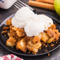 Apple brown betty on a black plate topped with scoops of vanilla ice cream, with a fork, cinnamon sticks, and a Honeycrisp apple in the background.