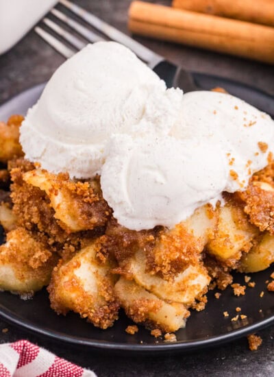 Apple brown betty on a black plate topped with scoops of vanilla ice cream, with a fork, cinnamon sticks, and a Honeycrisp apple in the background.