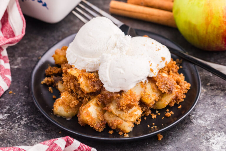Apple brown betty on a black plate topped with scoops of vanilla ice cream, with a fork, cinnamon sticks, and a Honeycrisp apple in the background.