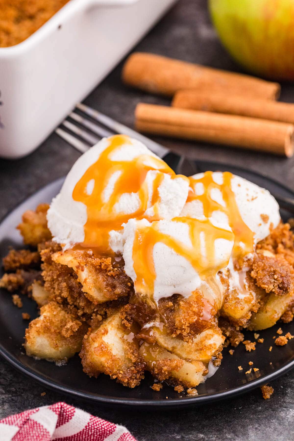 Apple brown betty on a black plate topped with vanilla ice cream and caramel sauce, with cinnamon sticks and a baking dish behind it.