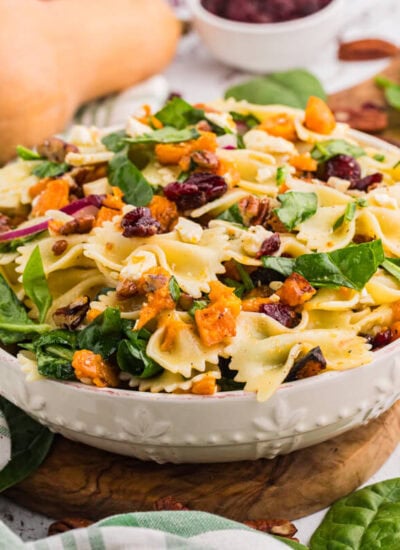 White bowl of fall pasta salad on a wooden board with bow tie pasta, roasted butternut squash, spinach, cranberries, pecans, and feta, with a whole butternut squash and a wooden spoon beside it.