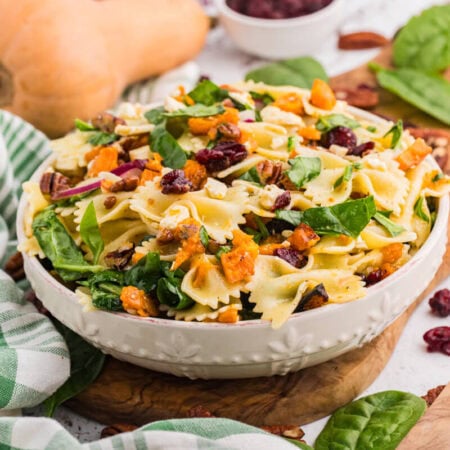 White bowl of fall pasta salad on a wooden board with bow tie pasta, roasted butternut squash, spinach, cranberries, pecans, and feta, with a whole butternut squash and a wooden spoon beside it.