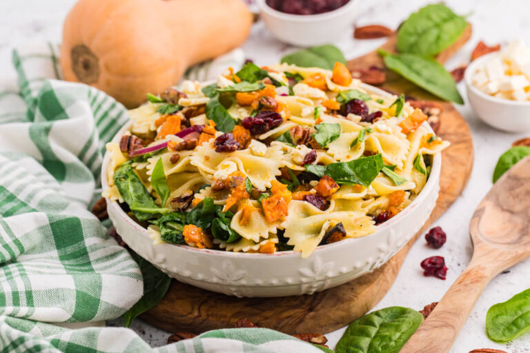 White bowl of fall pasta salad on a wooden board with bow tie pasta, roasted butternut squash, spinach, cranberries, pecans, and feta, with a whole butternut squash and a wooden spoon beside it.