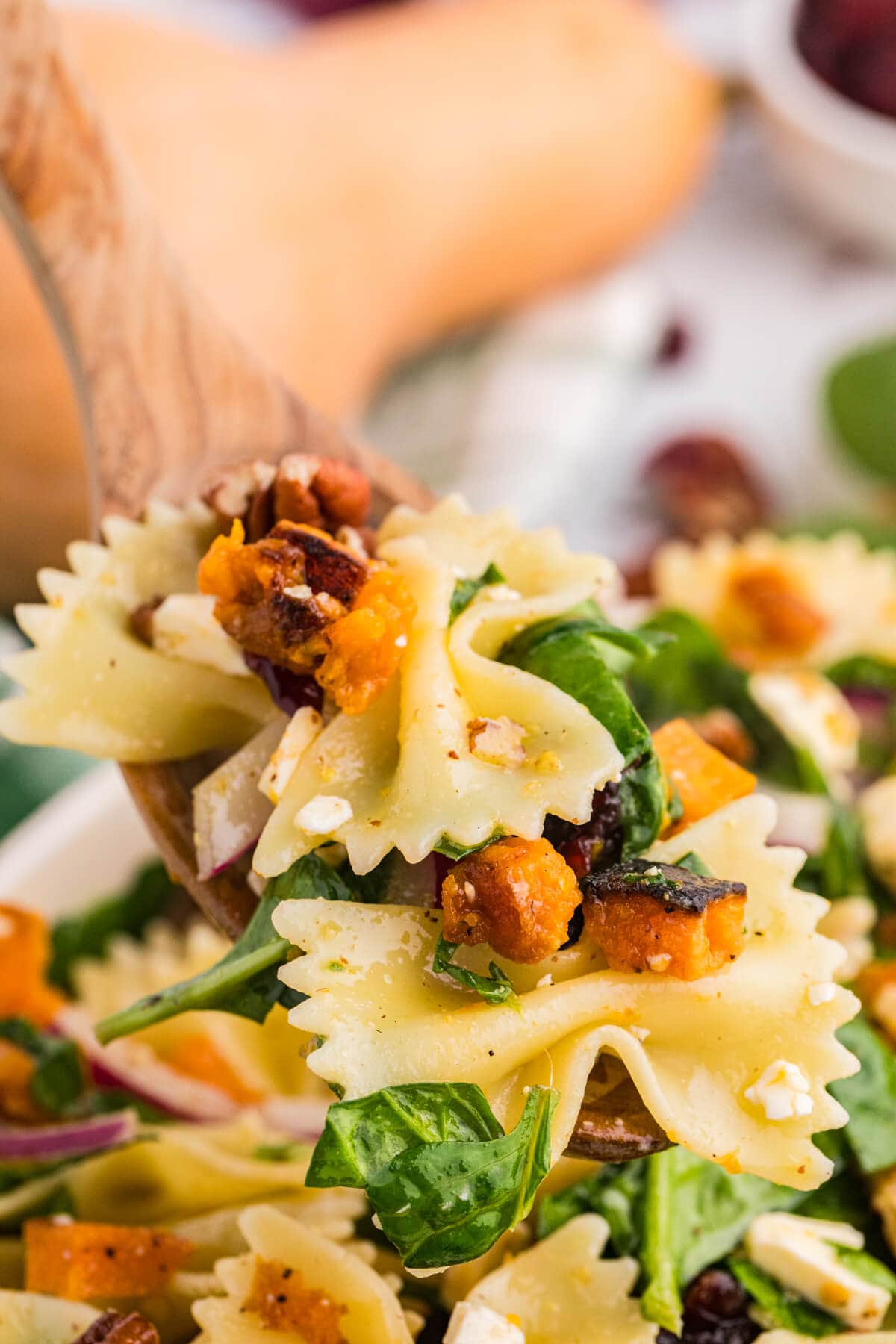 Wooden spoon lifting a forkful of fall pasta salad with bow tie pasta, roasted butternut squash, spinach, pecans, and feta.